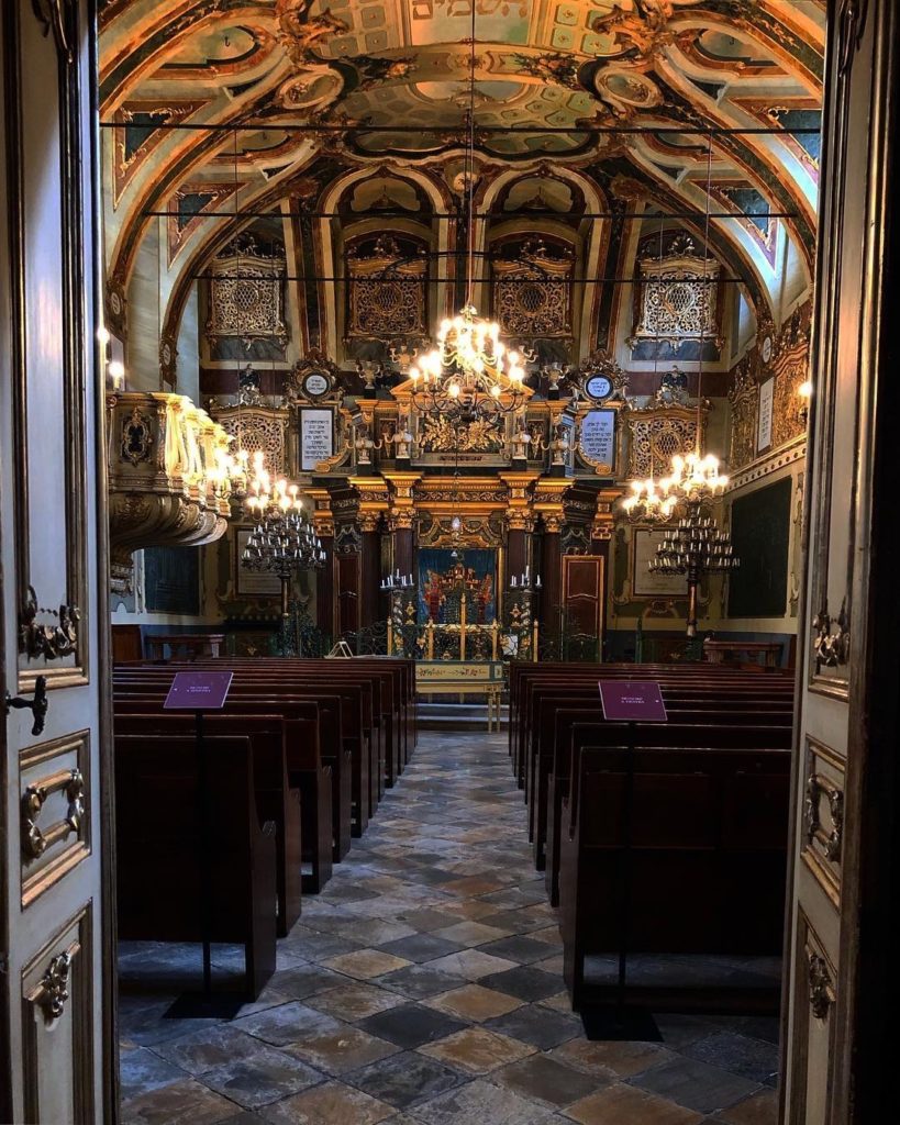 In foto l'entrata della sinagoga di casale monferrato vista dalla porta. immense le luci e le decorazioni dorate dell'altare, circondate da due file di banchi di legno scuro. Il sito fa parte dei tour virtuali di pinka group