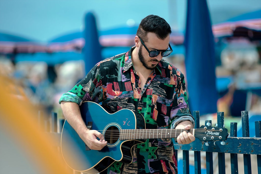 andrà tutto bene vemo. nella foto il cantante, sulla spiaggia, che indossa una camicia a fiori, gli occhiali da sole, con la chitarra a tracolla