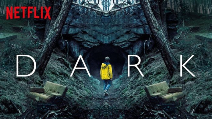 In foto la copertina della serie tv Dark, con sullo sfondo una grotta e due tronchi di albero. In centro tra le prime due lettere del titolo e le ultime due c'è il protagonista Jonas, di spalle con una giacca gialla che guarda verso la grotta.