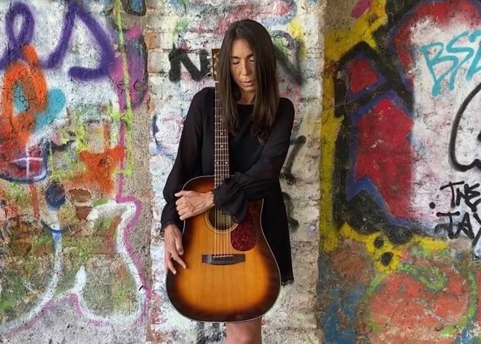 Patrizia Cirulli canta "C'est la vie" di Achille Lauro. Nella foto la cantante milanese, vestita di nero, con un chitarra tra le braccia, appoggiata a un muro dipinto con dei murales
