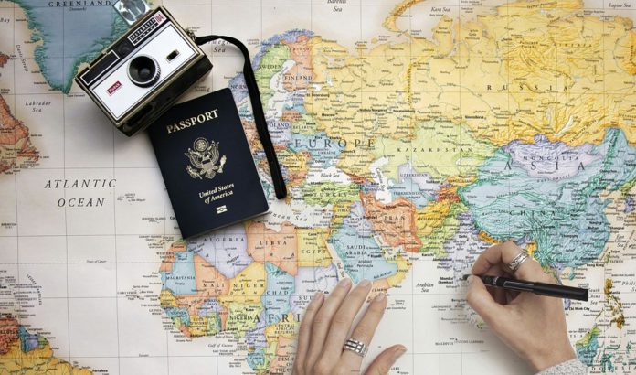 In foto una mappa del mondo, una macchina fotografica e un passaporto. Due mani pianificano i viaggi, una tiene stesa la cartina e l'altra tiene la biro tra le dita. Visibile anche l'Italia