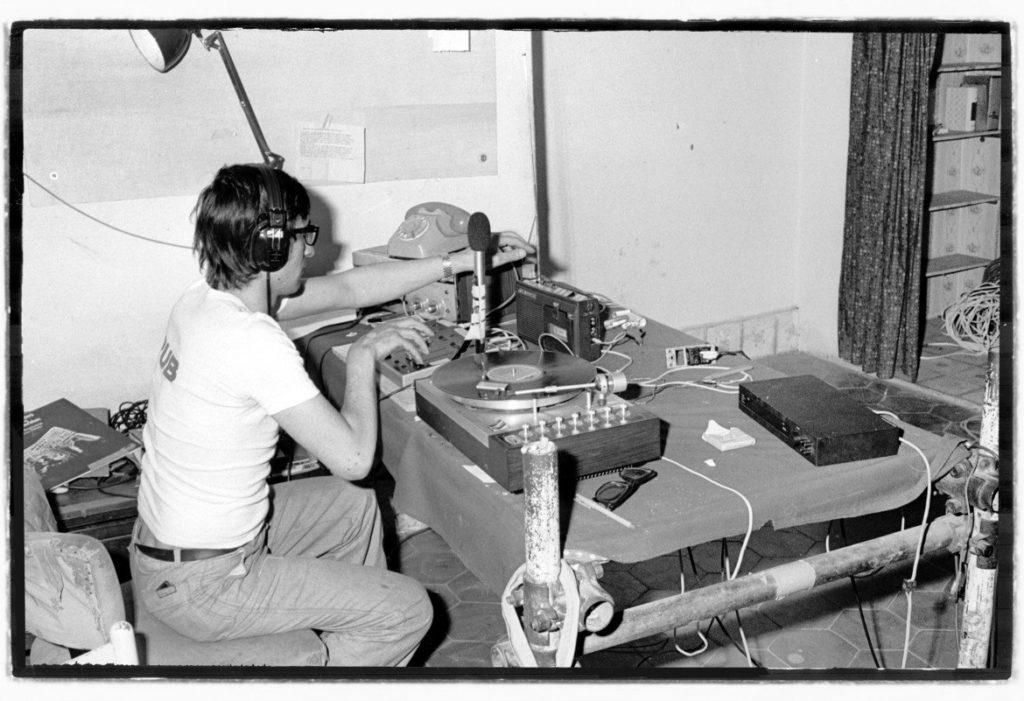 La storia della disco music: nella foto in bianco e nero, una radio libera anni settanta, con un ragazzo imegnato a trasmettere, un tavolo con sopra giradischi, microfono e registratore a cassette.