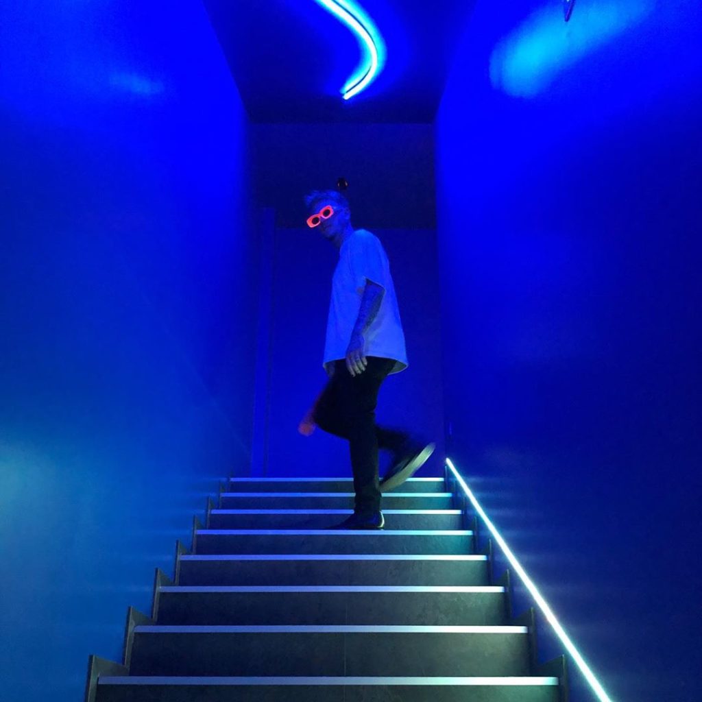Jack James feat. Brenda Carolina Lawrence rockin. Nella foto il rapper in piedi su una sclinata in mezzo a due muri, luci blu. Indossa occhiali da sole rossi, pantaloni neri e t-shirt bianca