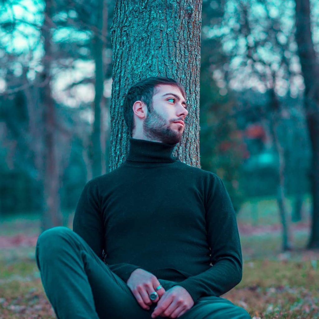 urano fratelli d'italia. appoggiato al tronco di un albero, il rapper, capelli corti e barba, indossa una maglia girocollo nera e pantaloni grigi