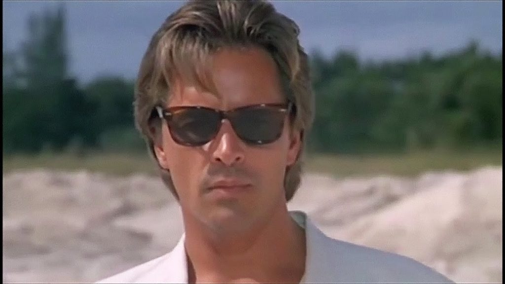 primo piano di don johnson sulla spiaggia, con addosso una giacca bianca e gli occhiali da sole