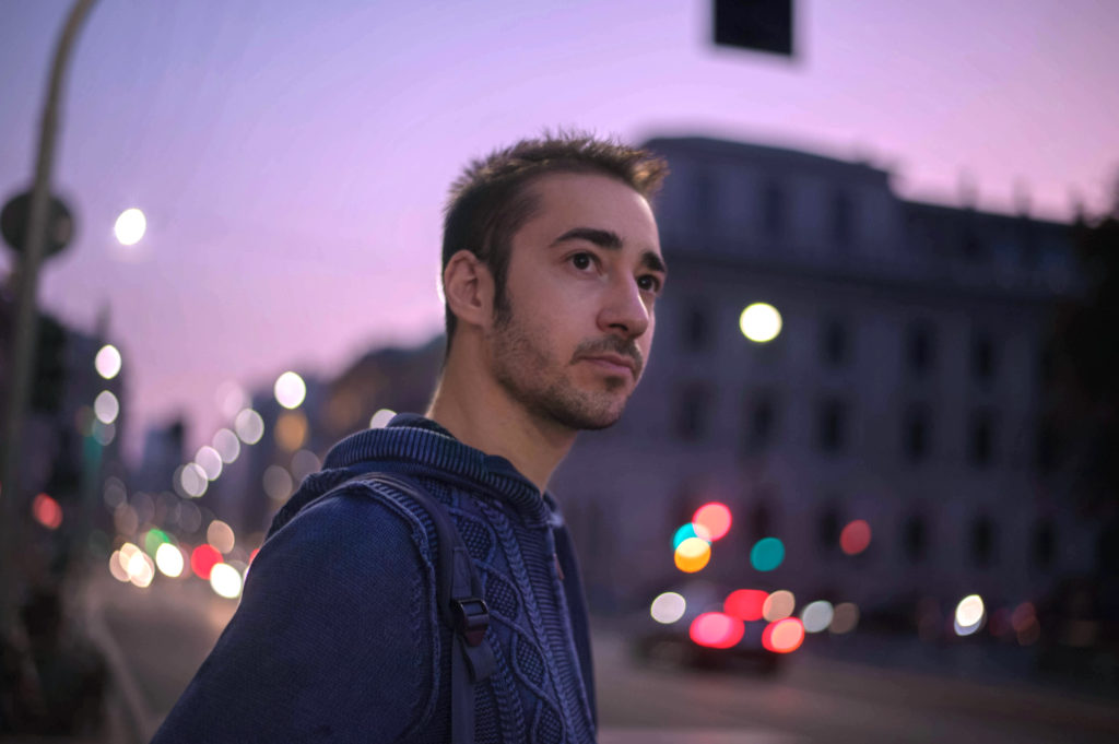 stefano bruno carlotta sfondo notturno e cittadino, in primo piano il cantante milanese, capelli corti, barba cortissima e felpa blu con cappuccio. di profilo