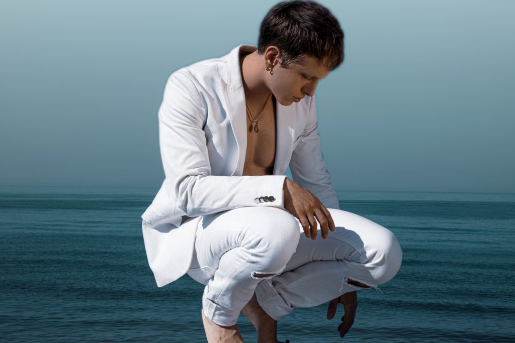 azzurro scontato giacomo eva - nella foto il giovane canttante accosciato sulla spiaggia, sullo sfondo il mare, vestito completamente di bianco