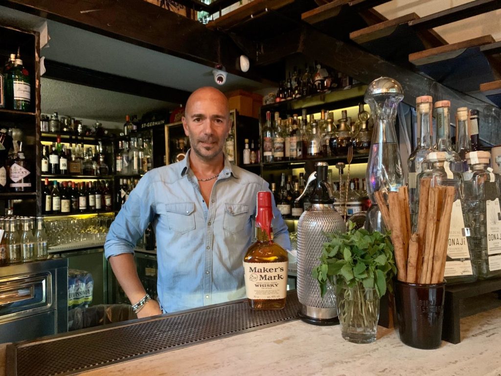 Shinig il drink e il barista che lo ha inventato dietro il bancone del suo bar, con tante bottiglie sugli scaffali