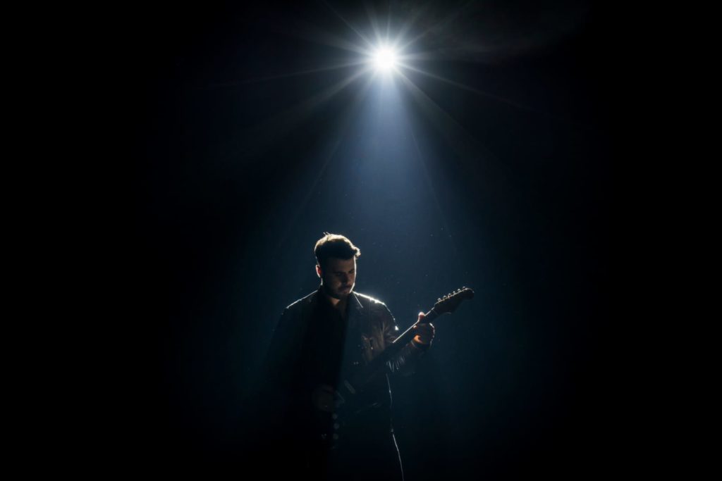 stefano delta fake - il cantante, chitarra a tracolla, al buoi su un palcoscenico, illuminato da una luce sopra la testa