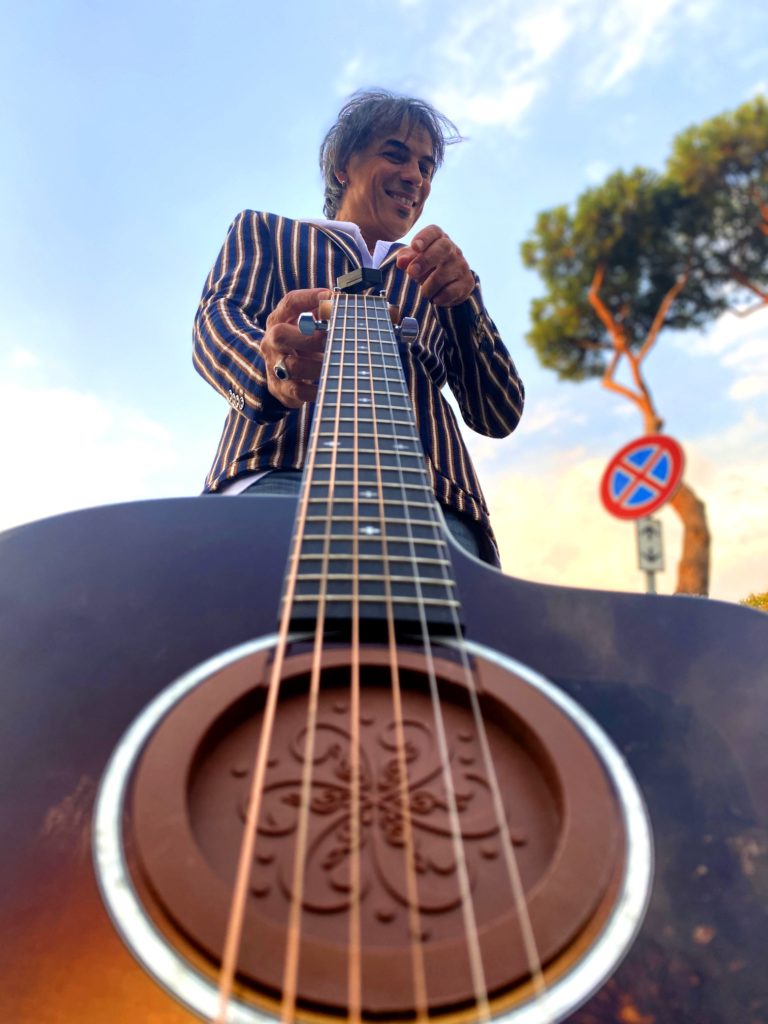 maurizzio fortini le donne finarmente - il cantautore romano giacca a righe che tiene una chitarra acustica per il manico, sullo sfondo il cielo e gli alberi