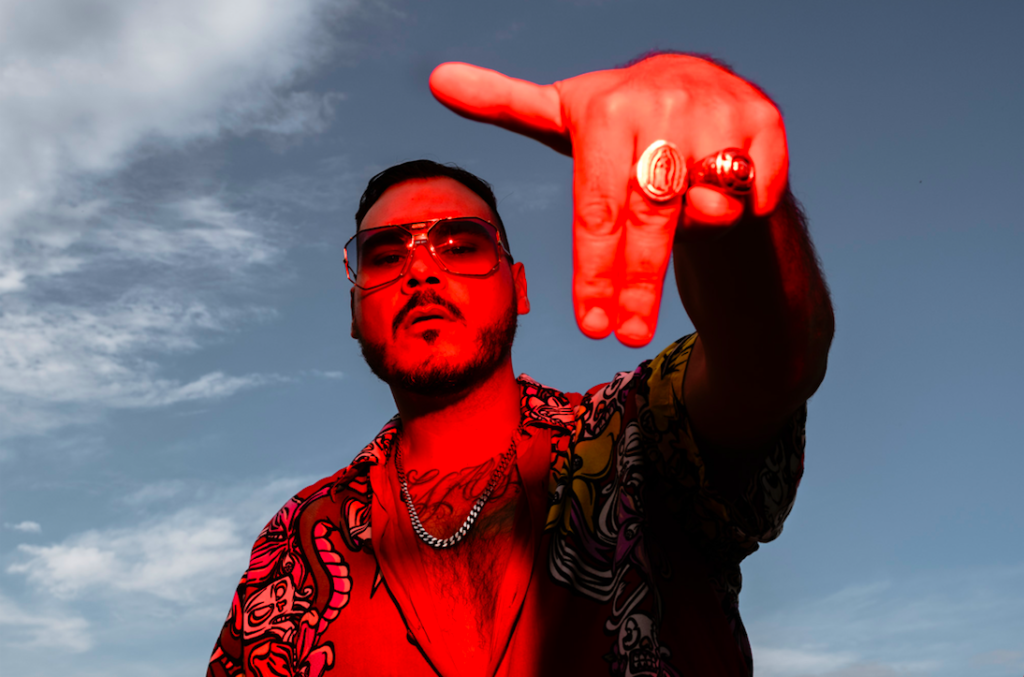 in foto il rapper Ensi in primo piano illuminato da una lue rossa, dietro lo sfondo del cielo. indica con due dita della mano verso il basso.