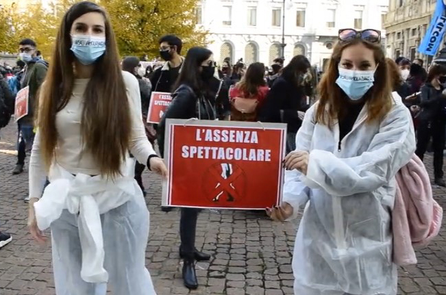 Proteste in piazza due ragazze vestite con tute bianche e mascherina tengono un cartello rosso con scritto 