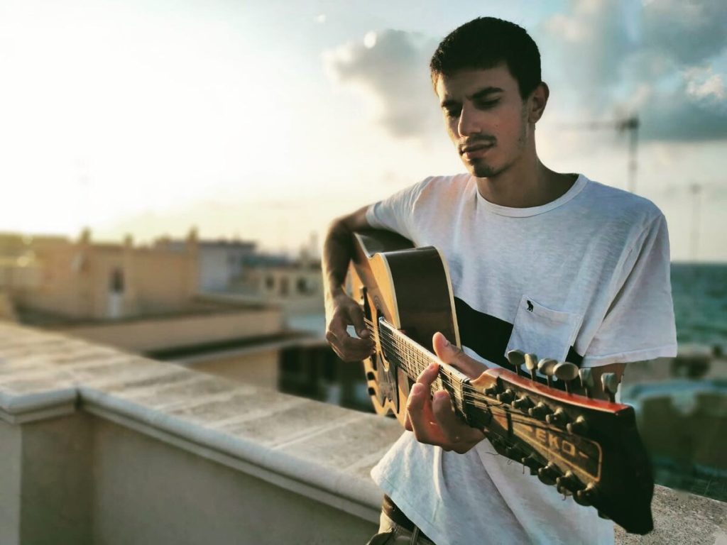 quando te ne vai - junior v - il giovane cantautore, seduto su un muretto con la chitarra acustica in mano, sullo sfondo i tetti della cittadina