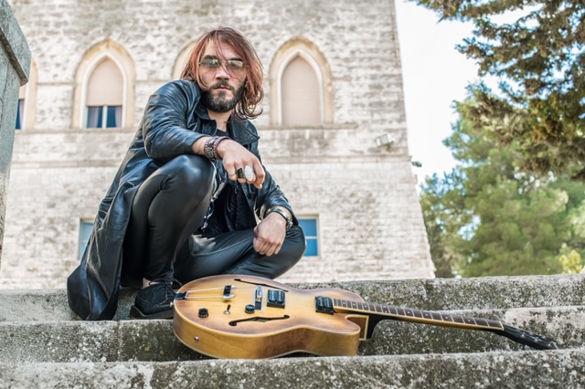 reverendo secret per te - il cantautore pantaloni e cappotto di pelle nera, capelli lunghi e occhiali da sole, inginocchiato su una scalinata, con ai piedi una chitarra elettrica di legno marrone