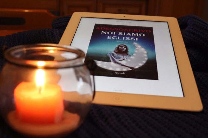 Noi siamo eclissi, romanzo, amore. In primo piano una candela di colore arancione accesa in un bicchiere di vetro. Dietro un tablet con la copertina del libro 