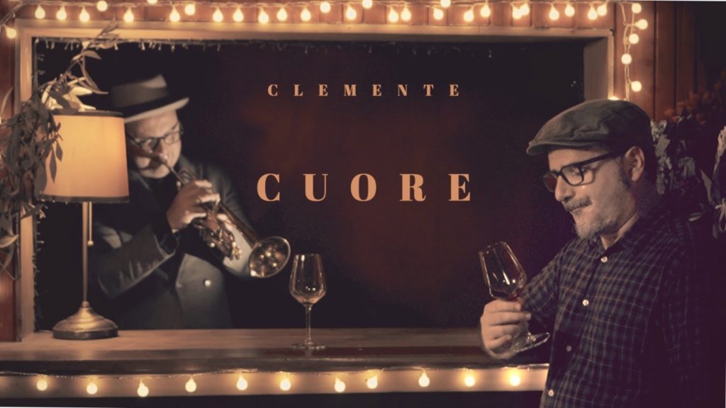 clemente cuore - il cantautore con un bicchiere di vino in mano, apppoggiato al bancone di un bar, dietro il bancone un musicista intento a suonare la tromba. 