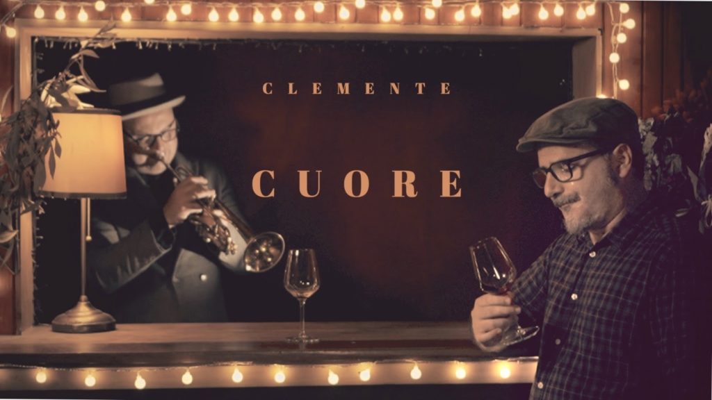 clemente cuore - il cantautore con un bicchiere di vino in mano, apppoggiato al bancone di un bar, dietro il bancone un musicista intento a suonare la tromba.