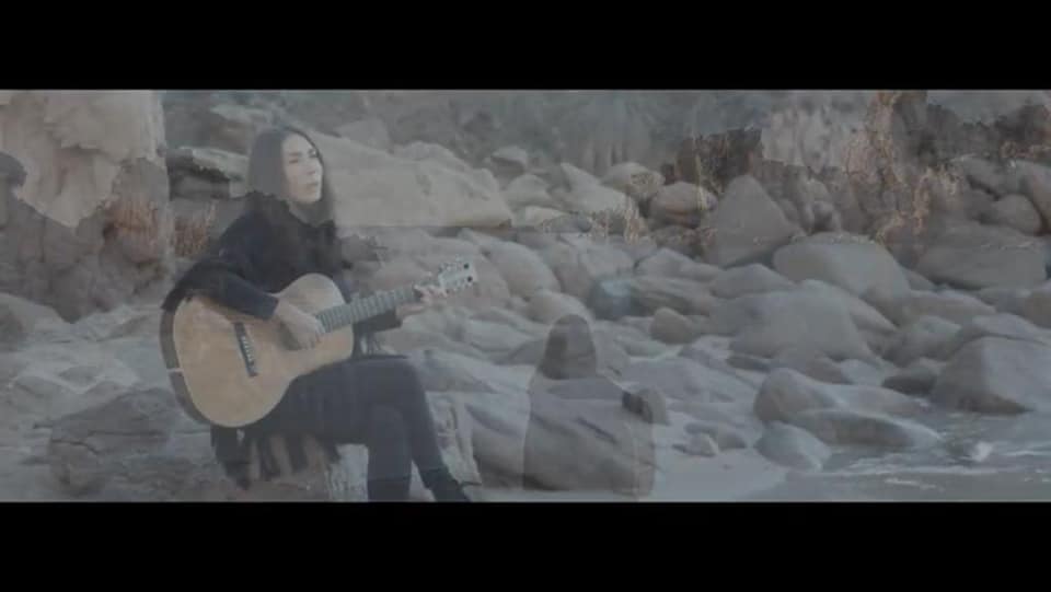 il mare immenso - patrizia cirulli sedura su uno scoglio, vestita di nero, con la chitarra appoggiata in grembo (frame del videoclip)