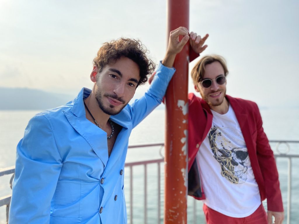 ossigeno (mi sei mancato) le tendenze - a sinistra francesco saibene, in giacca azzurra doppio petto, a destre daniele saibene, giacca rossa e sotto t-shirt bianca, appoggiati ad un palo. sullo sfondo il lago maggiore