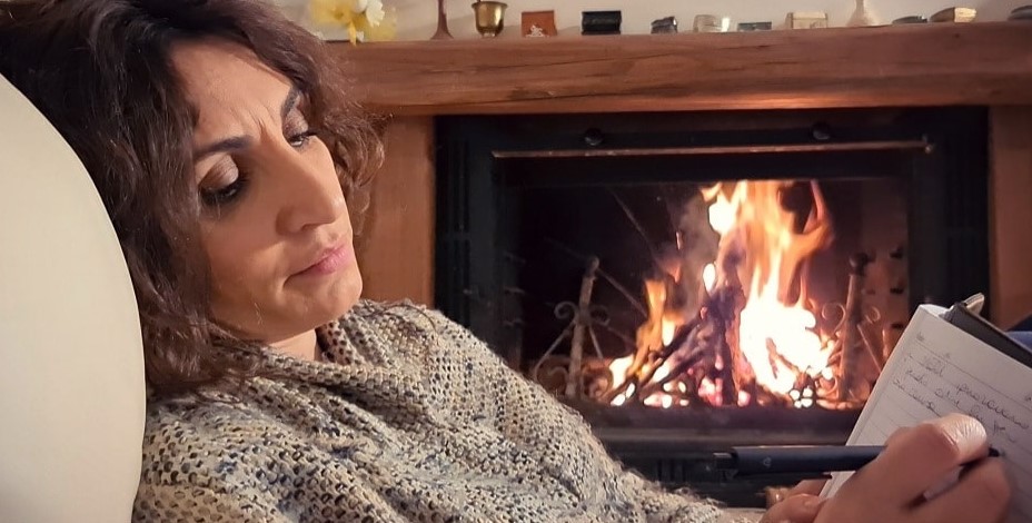 Paola Lavini di profilo seduta su una poltrona scrive su un tacquino e sullo sfondo un calino accesocon fiamme allegre