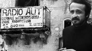 Peppino Impastato in un afoto in bianco e nero, con maglione nero e barba incolta. Dietro di lui un acasa e da un balcone un lenzuolo con scritto Radio Aut