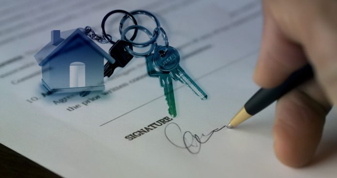 covid e mercato immobiliare - un contratto con. appoggiate sopra, delle chiavi con un portaachiiavi fatto a forma di casa e una mano che tiene una penna mentre firma il contratto