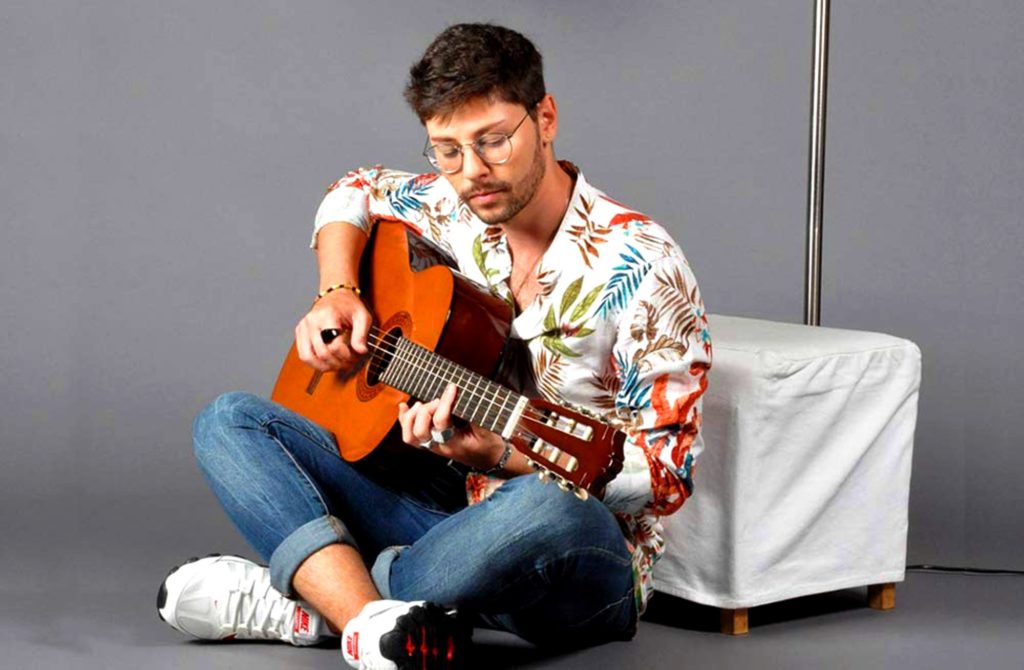 niccolò battisti seduto a gambe incrociate, jeans e camicia colorata, intento a suonare la chitarra