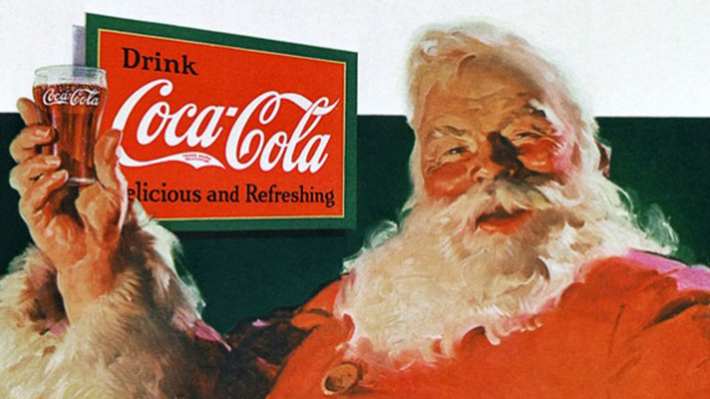 Curiosità riti e usanze di Natale - nella foto Babbo Natale vestito di rosso, con la barba bianca lunga, tiene in mano un bicchiere di coca cola e dietro di lui il pannello pubblicitario con la scritta "Coca Cola"