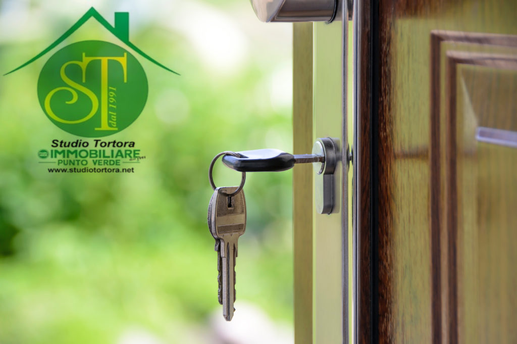 comprare e vendere casa, nella foto un primo piano di un mazzo di chiavi inserito nella toppa di una porta e sullo sfondo sfocato un giardino. A sinistra in alto, il logo dell'imoobiliare Punto Verde Studio tortora con un pallino verde e sopra un tetto e nel pallino le iniziali "ST"