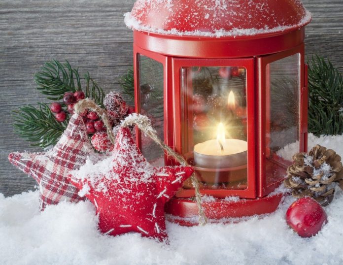 Una candela accesa in una lanterna rossa appoggiata sulla neve e intorno due stelle, un abianca e una dorata, dell'agrifoglio con delle bacche rosse, una pgna e una mela