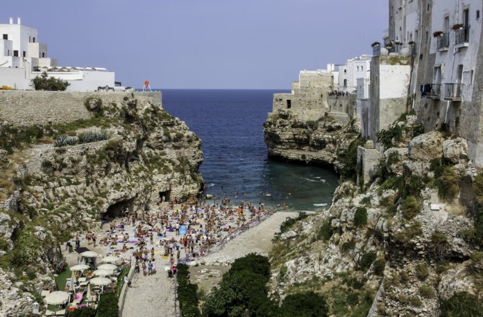 Polignano a Mare, opere, mare. IL paesaggio presenta un cielo con i colori del tramonto. Il mare avvolto dagli scogli e da case basse.