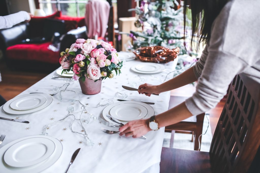 Una donna prepara la tavola per il pranzo di Natale, con piatti bianchi e posate argentate,  a centro tavola un vaso di fiori e al fondo una decorazione natalizia