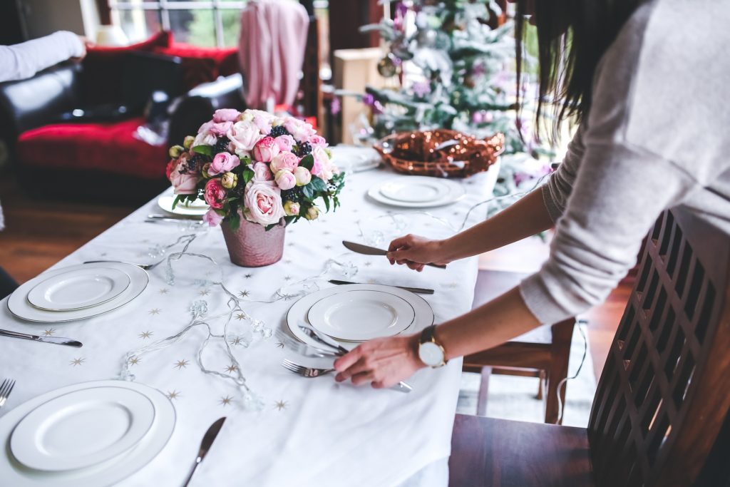 Una donna prepara la tavola per il pranzo di Natale, con piatti bianchi e posate argentate, a centro tavola un vaso di fiori e al fondo una decorazione natalizia
