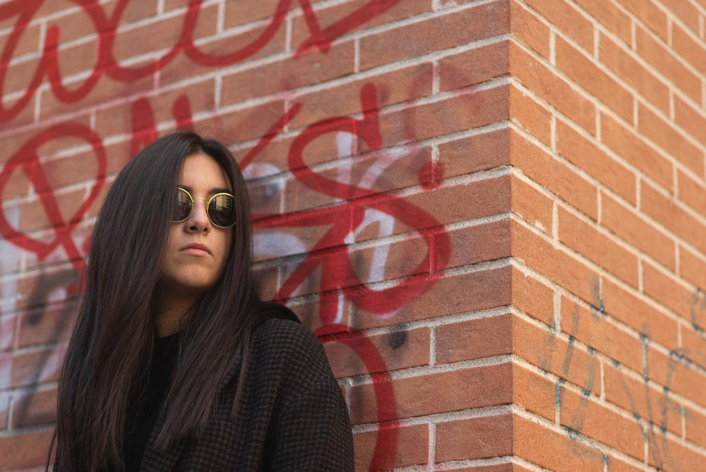 lena A, capelli lunghi neri, giacca nera, occhiali da sole, appoggiata a un muro di mattoni