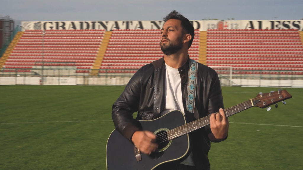 il discorso del capitano - leone, t-shirt bianca e giacca di pelle, chitarra nera a tracolle. sullo sfondo la gradinata dello stadio moccagatta di alessandria