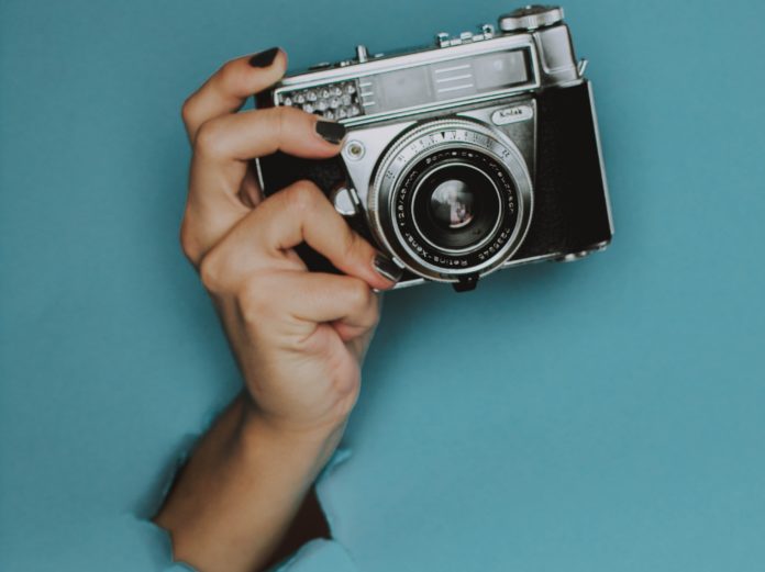 una macchina fotografica tenuta da una mano ceh sbuca da uno sfondo azzurro