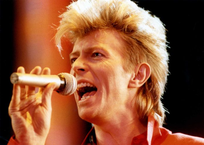 David Bowie mentre canta con microfono in mano capelli biondi e camicia rossa