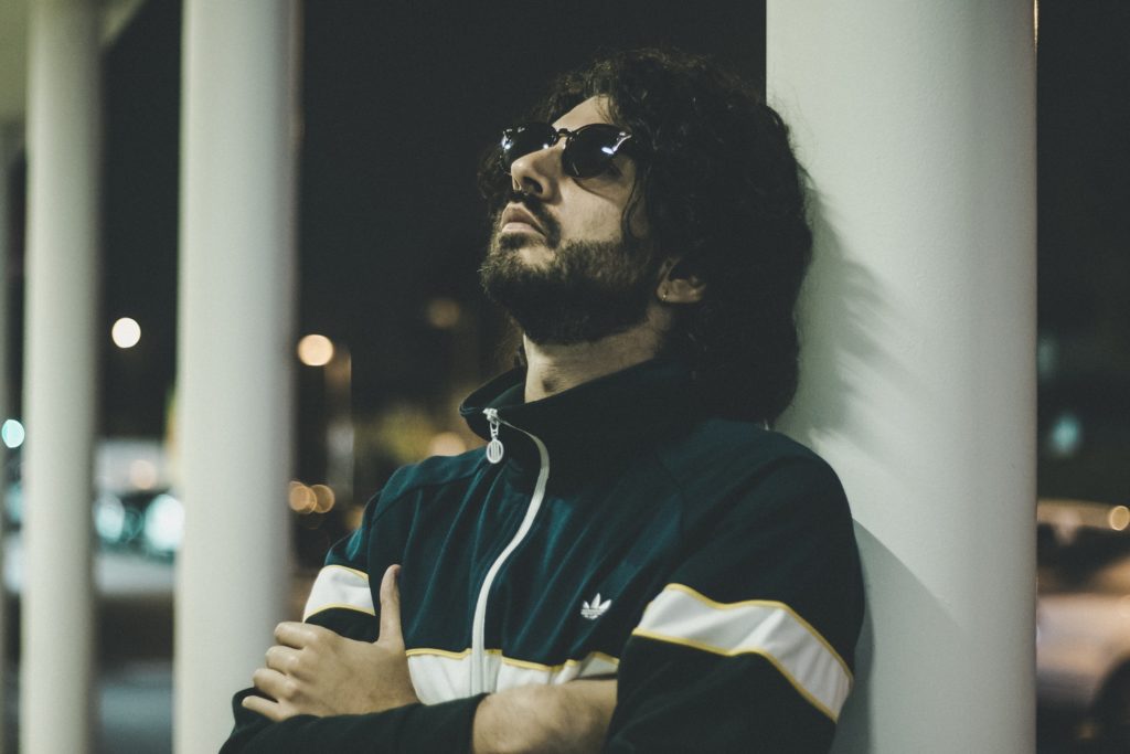 Mosè Santamaria appoggiato a un muro bianco, capelli lunghi scuri, barba e occhiali da sole, indossa una tuta blu adidas con banda bianca