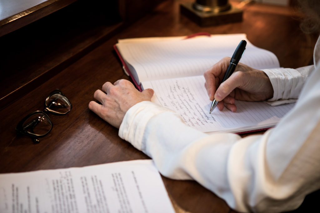 Camilla Ugolini Mecca, seduta ad una scrivania di legno scrive con una penna sul quaderno, occhiali riposti vicino alla mano sinistra 
