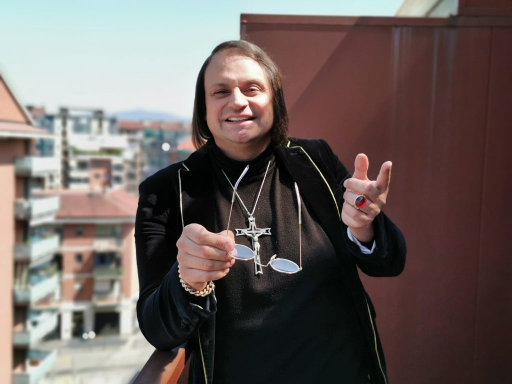 Italia al tempo del Covid- nella foto Maurizio Scandurra, appoggiato alla ringhiera di un balcone dal quale si vede uno scorcio di panorama fatto di palazzi, sorride e tiene in mano degli occhiali. Maurizio è vestito di nero con un vistoso crocifisso al collo