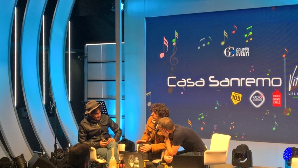 In foto Casa Sanremo, area in cui i cantanti in gara vengono intervistati. anno 2018, Ermal Meta e Fabrizio Moro sono seduti e intervistati dal giornalista. da sinistra Moro ha un cappello e un giubbotto di pelle nero, meta una giacca color renna, ultimo è il giornalista con una maglietta nera, appoggia la testa sulla mano. Sullo sfondo il logo di Casa Sanremo, tutto blu e azzurro  