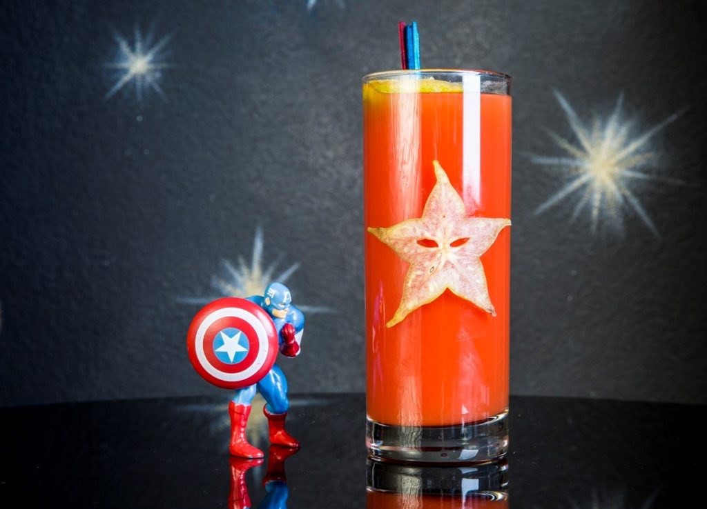 Capitan America con la tuta blu e rossa, con lo scudo rosso e bianco con una stella biaca al centro vicino ad un enorme bicchiere con una stella e dentro un drink di colore rosso 