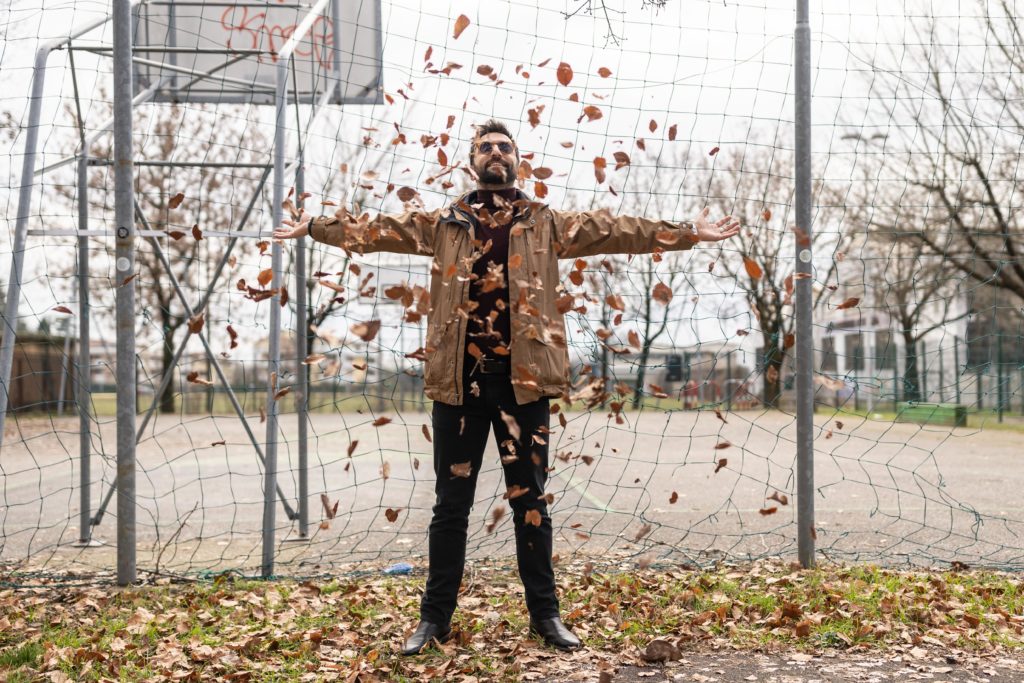 delorean - riccardo inge pantaloni scuri e giaccone beige, una pioggia di foglie che lo circonda, dietro la rete di un giardino, sullo sfondo un campo da basket all'aperto