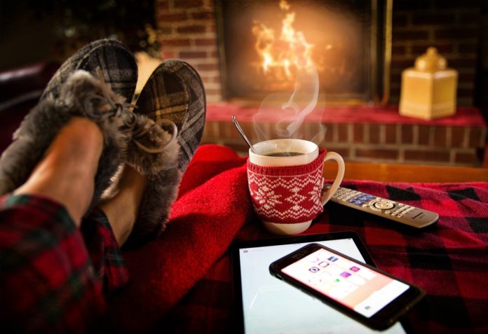 relax e pace l'immagine di due piedi con delle pantofole, appoggiati su un tavolino con affianco una tazza dfumante, un telecomando, un telefonino e sullo sfondo un camino acceso