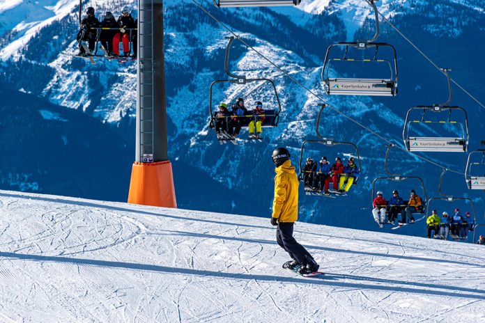 sci e sciatori au una seggiovia in montagna con sotto di loro una discesa di neve e un ragazzo con giubbotto giallo e casco nero che fa snowboarding