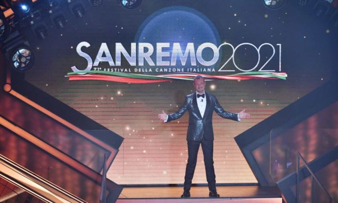 in foto Amadeus entra all'Ariston, allarga le braccia, indossa uno smoking grigio con camicia bianca, dietro la scritta Sanremo 2021 del logo