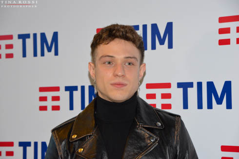 Irama in una foto del Festival di Sanremo del 2019 con giacchino di pelle beige, con capelli ricci corti biondi e sullo sfondo lo sposnor della Tim