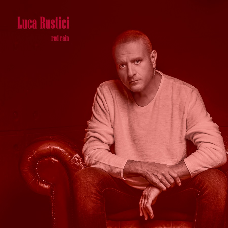 red rain luca rustici - il musicista seduto su una poltrona di pelle rossa, indossa una maglia bianca. lo sfondo è rosso scuro, come la scritta con il titolo e l'interprete, in alto a sinistra