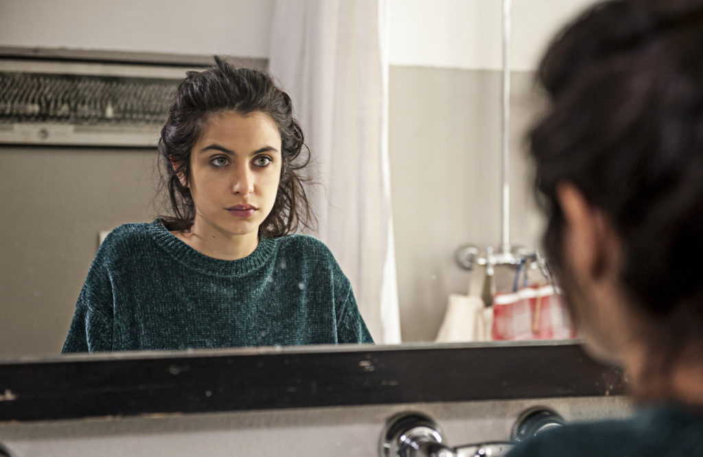 Una ragazza adolescente davanti allo specchio si guarda seria, ha lunghi capelli neri, una maglia verde e appoggia entrambi le mani su un ripiano