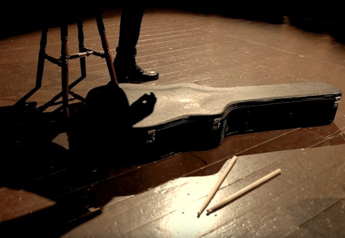 Una storia al presente - Dodi Battaglia - nella foto sul palcoscenico di legno di un teatro, si vede l'ombra di Dodi Battaglia, le gambe di uno sgabello e appoggiata sul pavimento la custodia della chitarra e di fianco, due bacchette da batteria